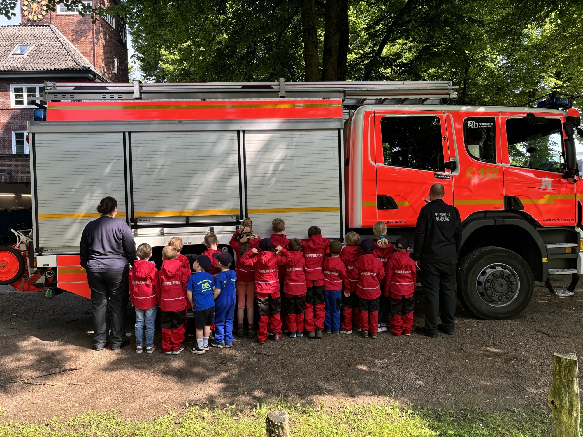 Kinder der Minifeuerwehr Ohlstedt in roten Einsatzjacken betrachten gemeinsam mit Betreuern ein Feuerwehrfahrzeug